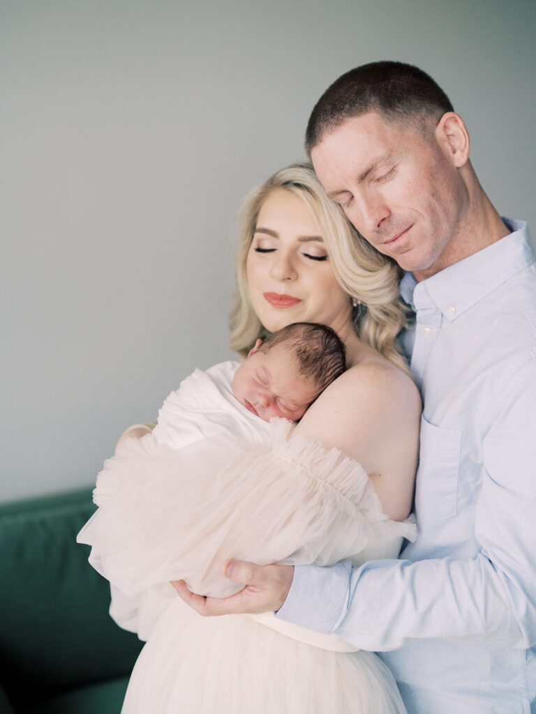 A Mother And Father Lean Into One Another While Holding Their Newborn Baby During Their Bethesda, Maryland Newborn Session.