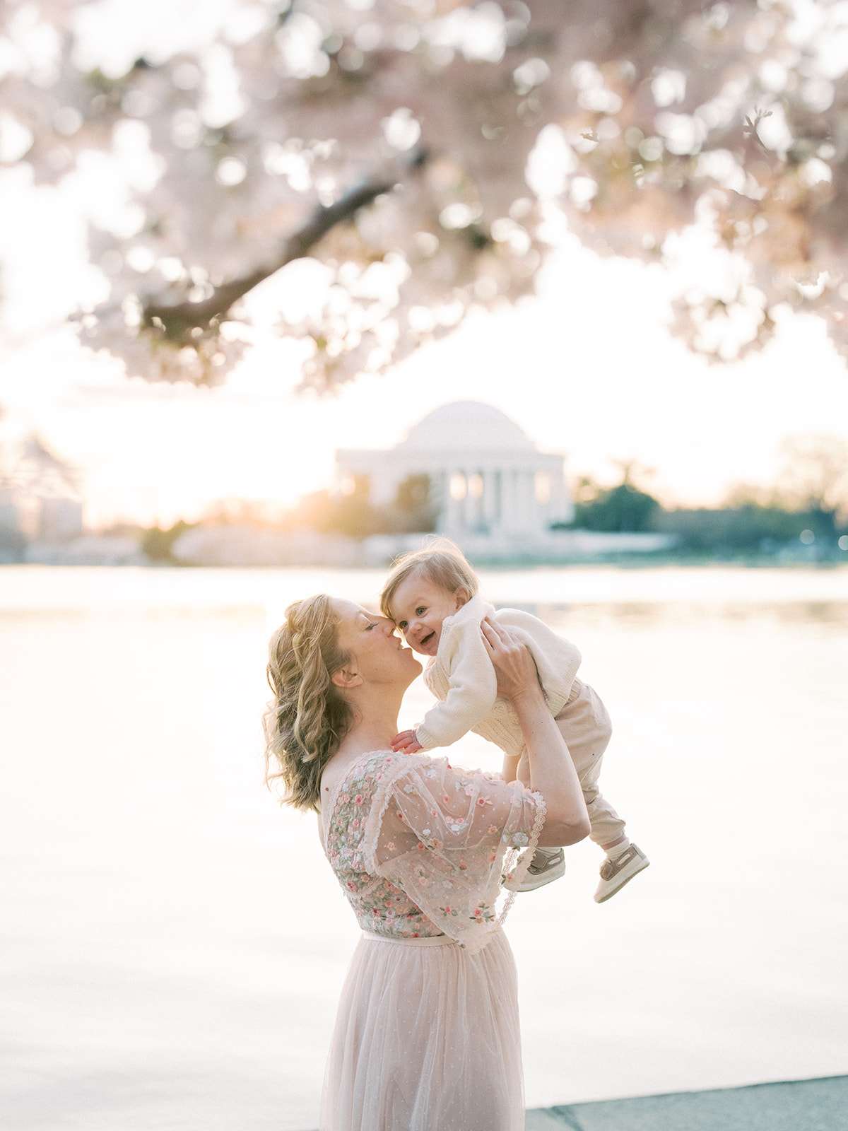 A Blonde Mother In A Pink Dress Stands Along The Dc Cherry Blossoms At The Tidal Basin Holding Up Her Young Son.