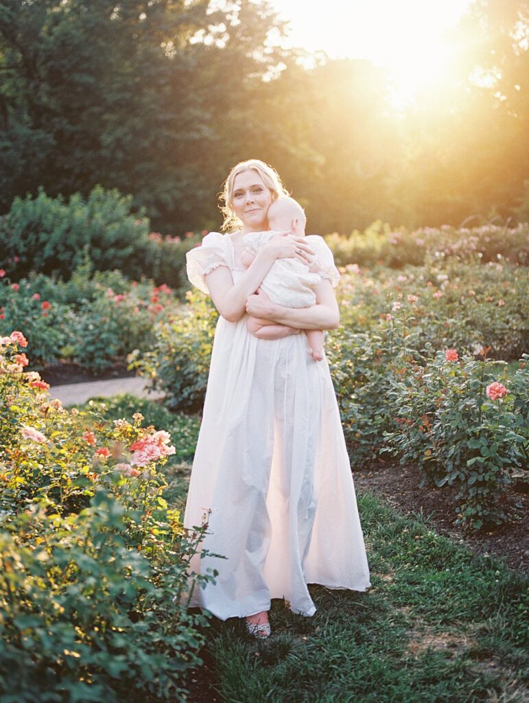 A Blonde Mother Stands In Bon Air Rose Garden At Sunset Holding Her Baby During Their Arlington Family Photos.