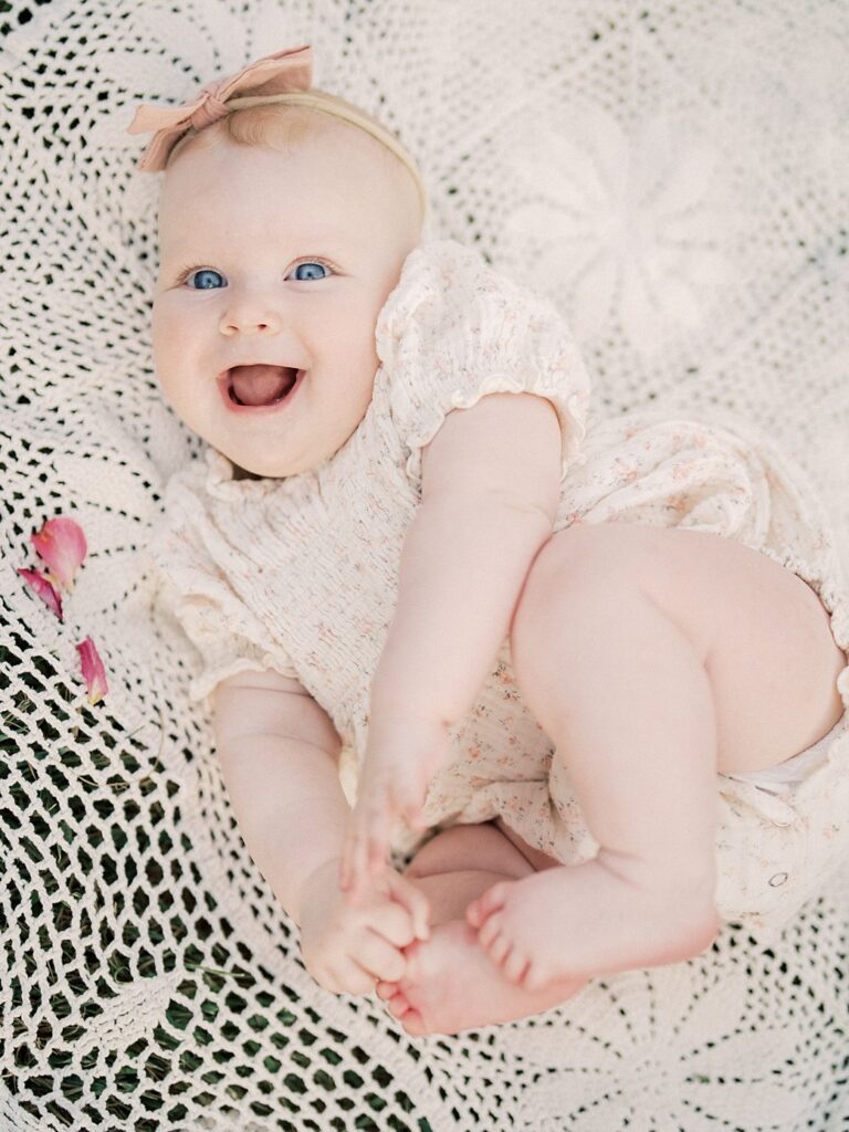 A Baby Girl Lays On A Crotchet Blanket With Rose Petals Smiling.