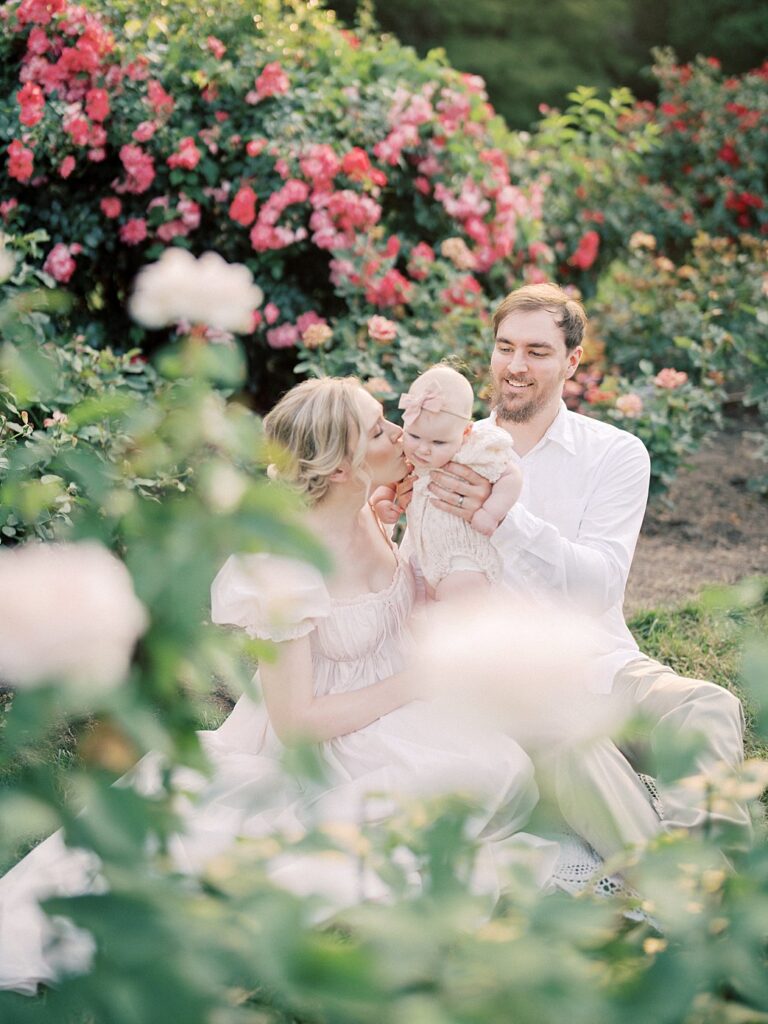 A Father Holds His Baby Over To His Wife Who Kisses The Baby As They Sit In A Garden During Their Arlington Family Photos.