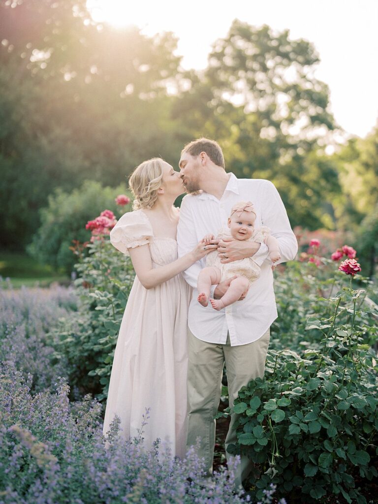 A Mother And Father Kiss As They Hold Their Baby In Bon Air Rose Garden.