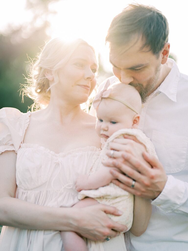 A Father Leans Down To Kiss His Baby's Head Who Is Held By His Wife.