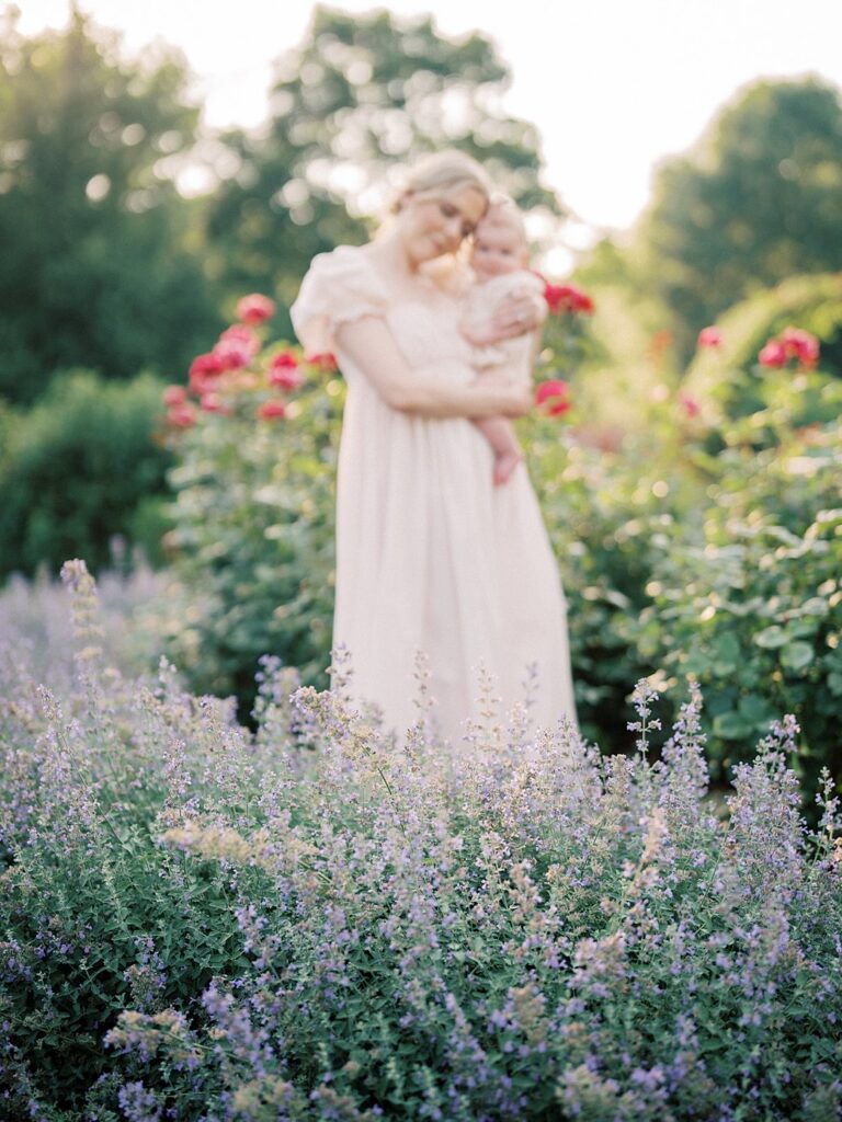 A Mother Holds Her Baby Out Of Focus As They Stand In A Garden With Roses And Purple Flowers.