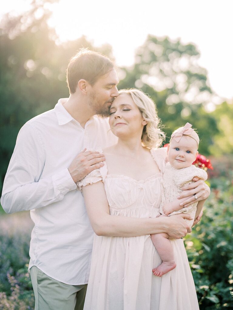 A Father Leans Over To Kiss His Wife Who Has Her Eyes Closed And Holds Her Daughter During Their Arlington Family Photos.