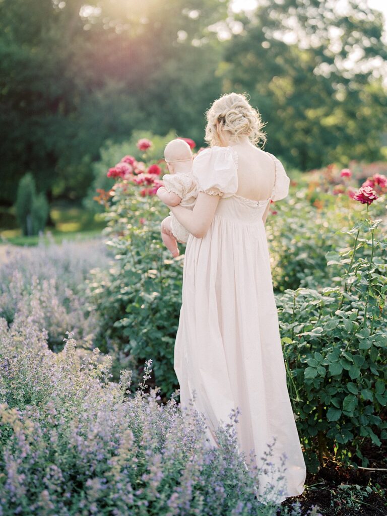 A Mother In A Light Pink Dress Walks Through A Garden Holding Her Daughter.