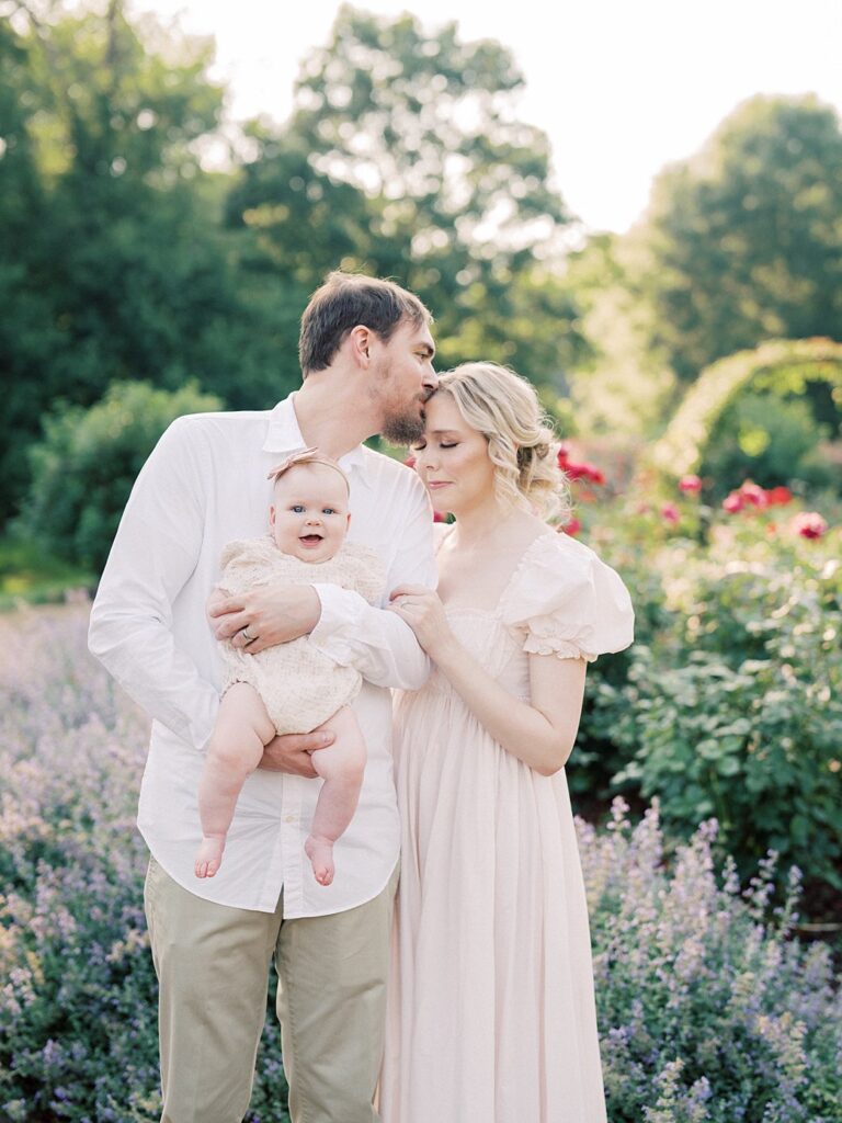 A Father Leans Over To Kiss His Wife's Forehead While Holding Their Baby In A Garden During Their Arlington Family Photos.