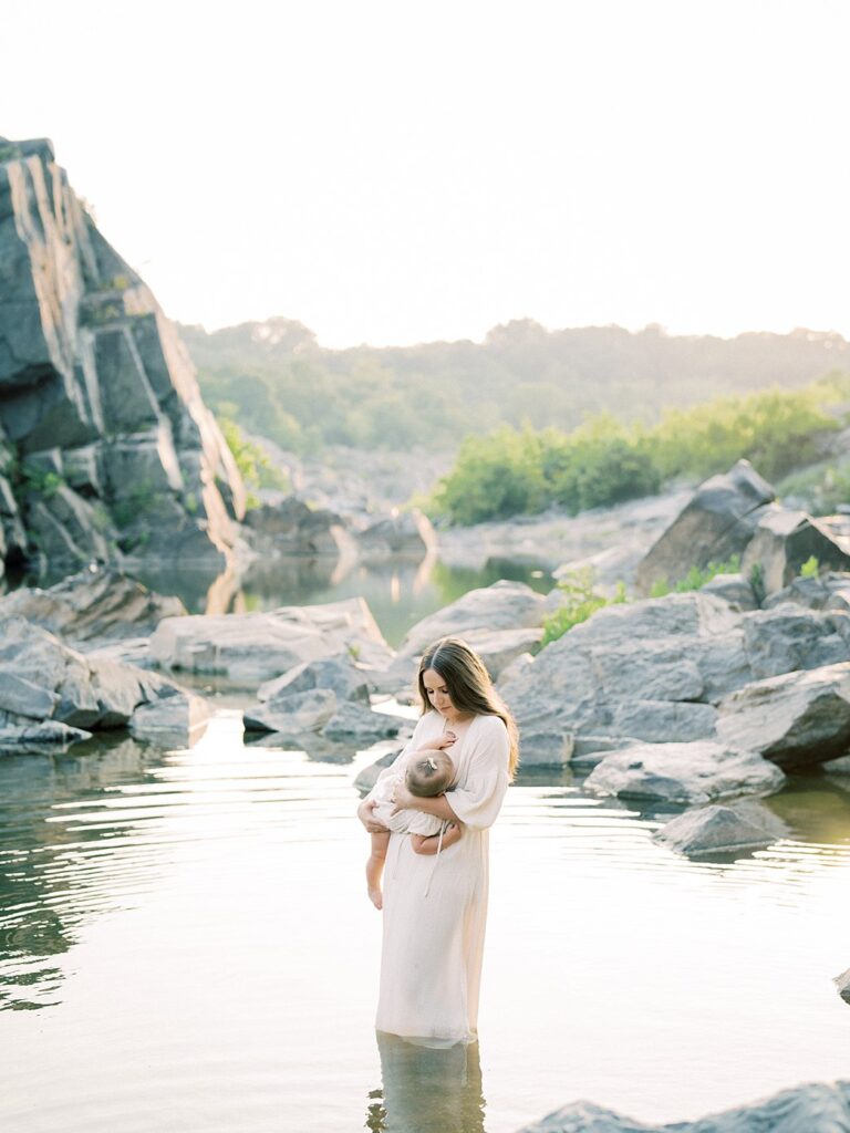 Mother Stands In The Water Nursing Her Baby At Great Falls Park During Their Family Photos.