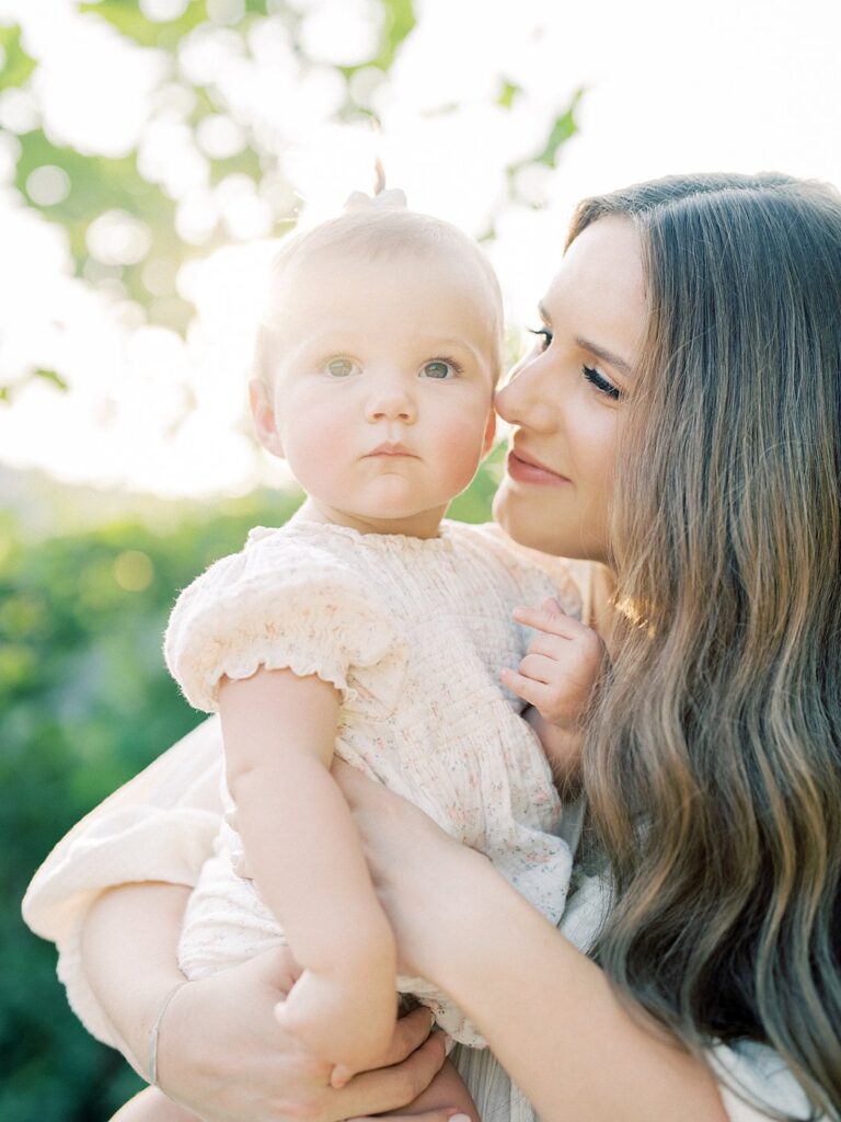 Mother With Long Brown Hair Holds Her One Year Old Daughter Up To Her Face As Her Daughter Gazes Away.