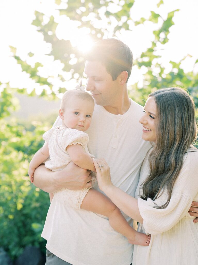 Father And Mother Hold Their Baby Who Looks At The Camera With A Soft Smile.