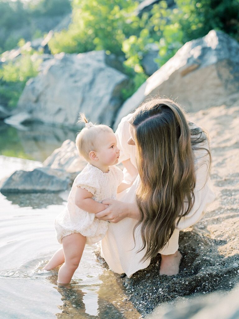 A Mother With Long Brown Hair Leans Down To Look At Her One Year Old Daughter In The Water.