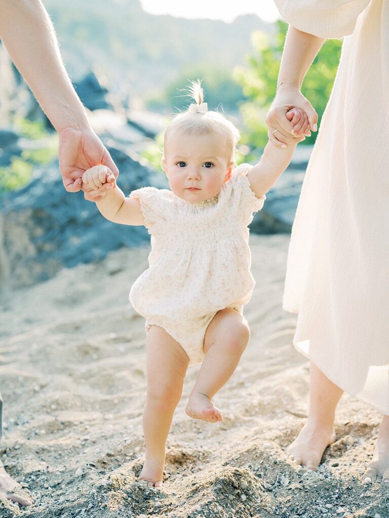 A One Year Old Year Tentatively Walks On A Sandy Beach While Holding Her Mother And Father's Hands.