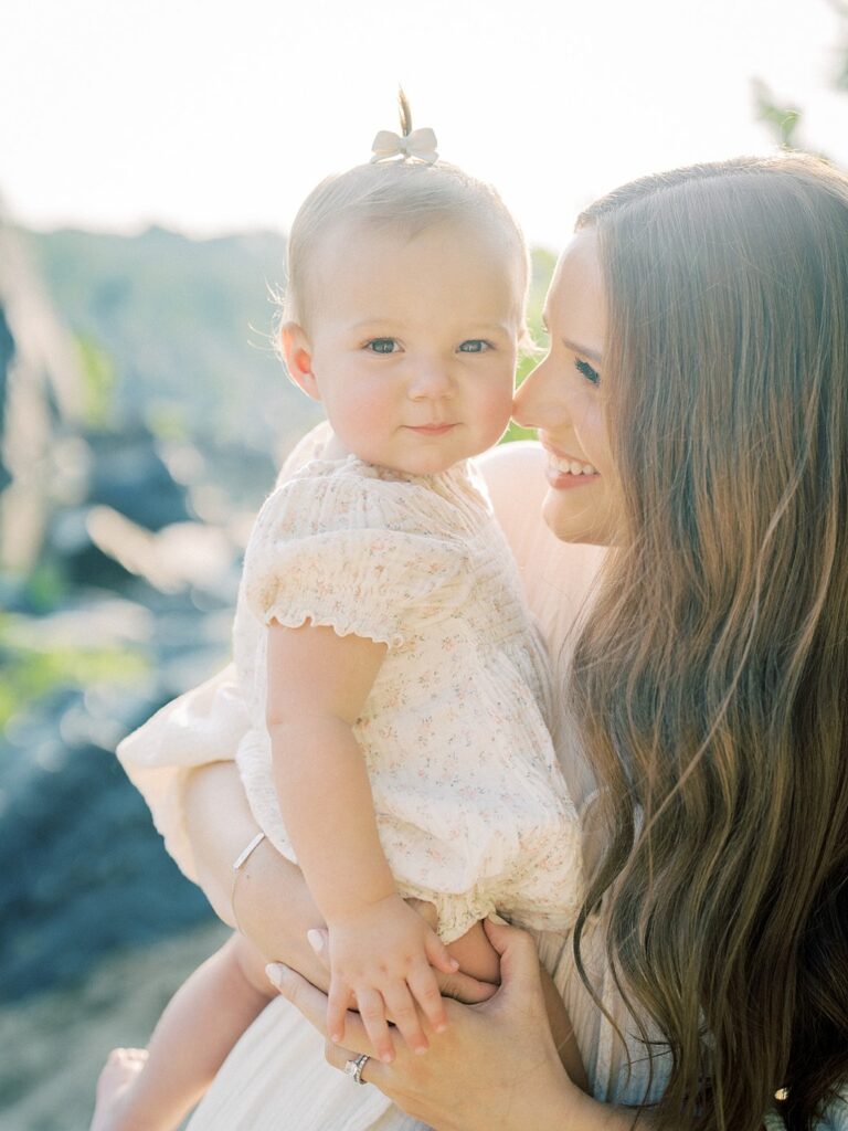 A Mother With Long Brown Hair Leans Into Her One Year Old Daughter While Holding Her At Sunset.