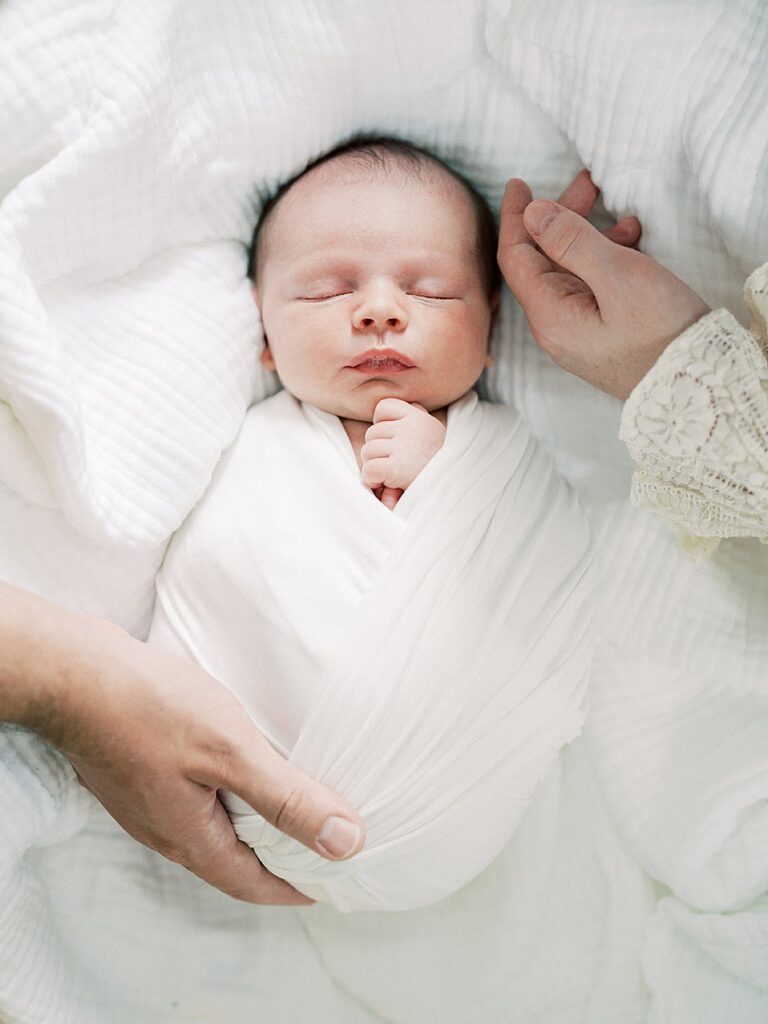 A Newborn Baby Sleeps Peacefully While Swaddled As His Mother And Father Each Lay A Hand On Him During Their Olney Newborn Photos.