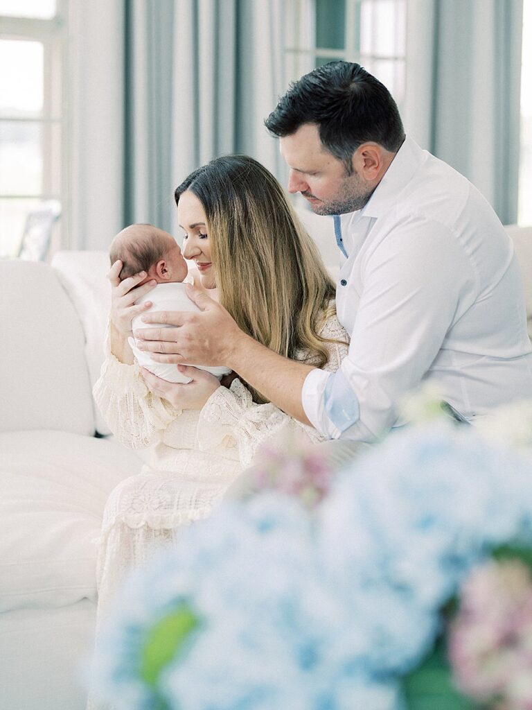 A Mother Holds Her Newborn Baby Up To Her Nose As She Sits With Her Husband In Their Living Room With Hydrangreas.