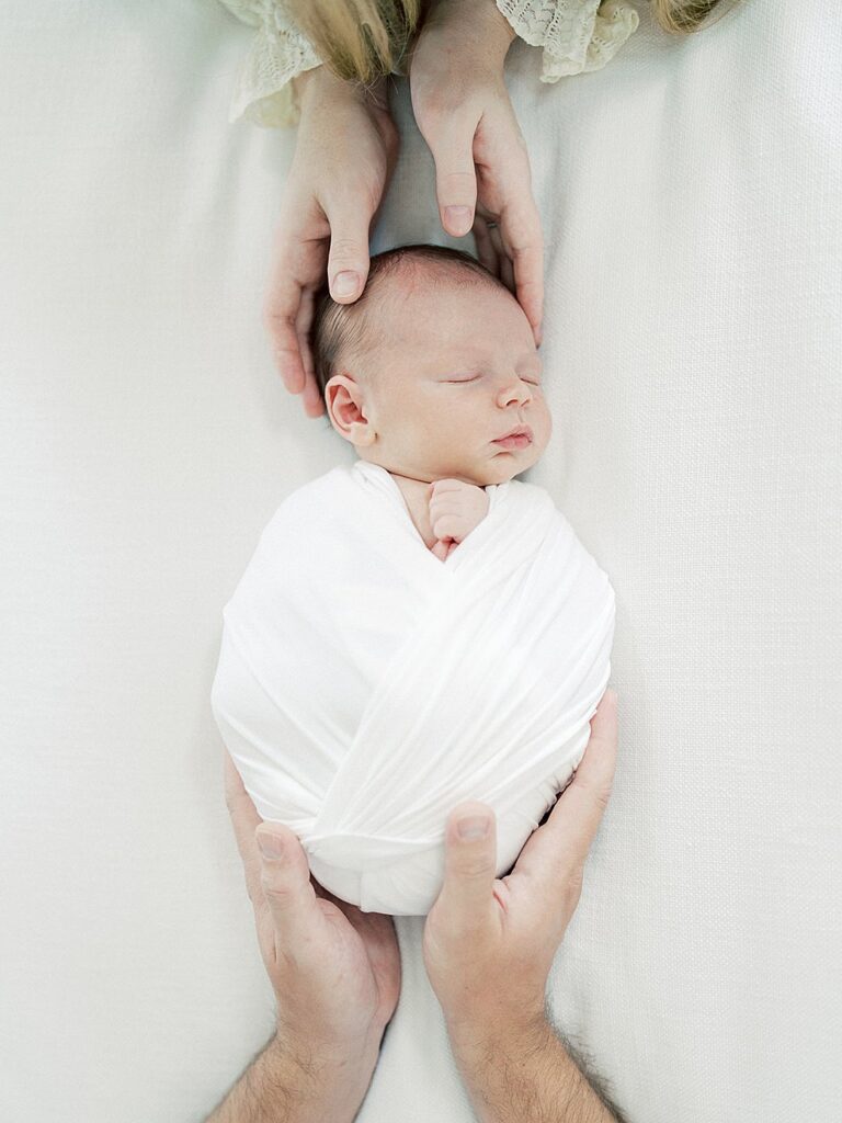 A View From Above As A Newborn Swaddled In White Is Held At The Head And Bottom By His Parents.