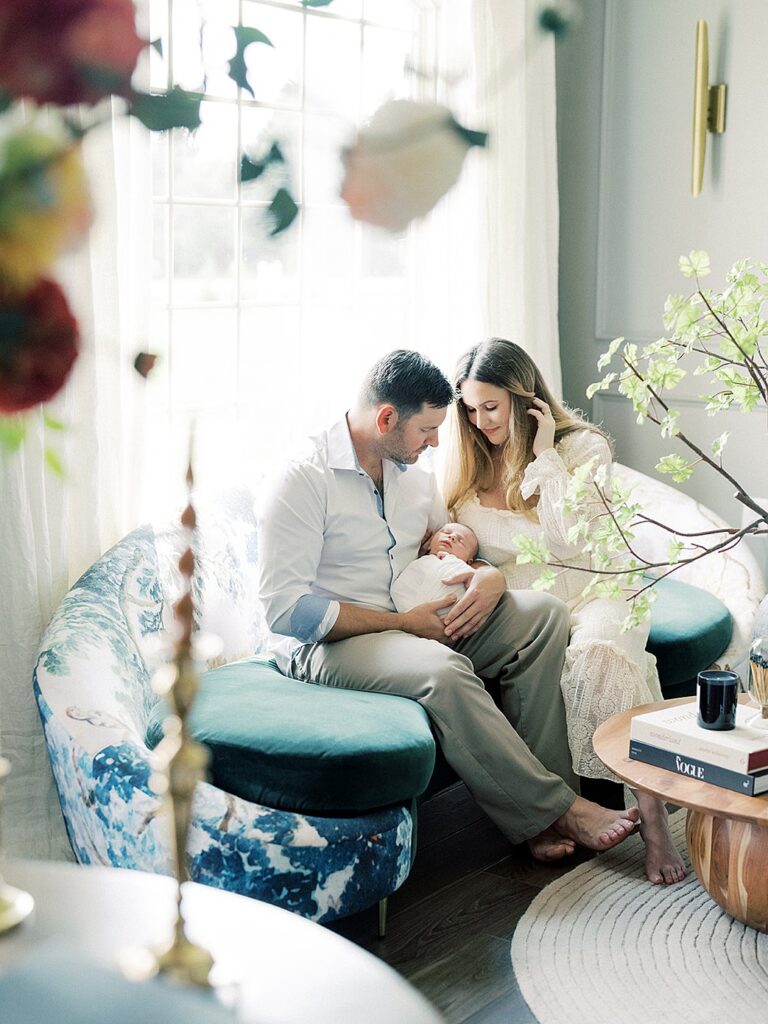 A Mother Leans Into Her Husband As He Holds Their Newborn Baby On Their Green Couch In Their Living Room During Their Olney Newborn Photos.