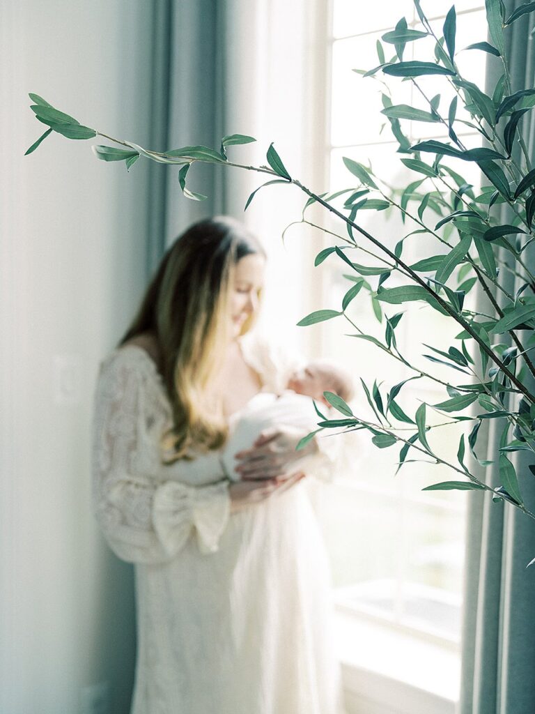 A Mother Holds Her Baby Boy Out Of Focus By A Window.