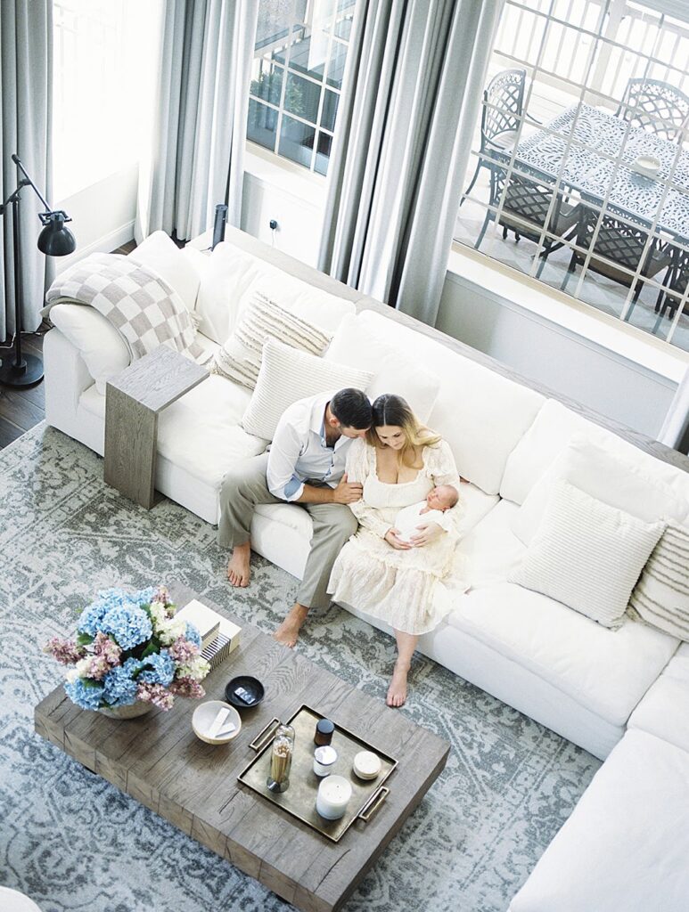 A View From Above A Living Room Of New Parents Holding Their Baby On The Couch.