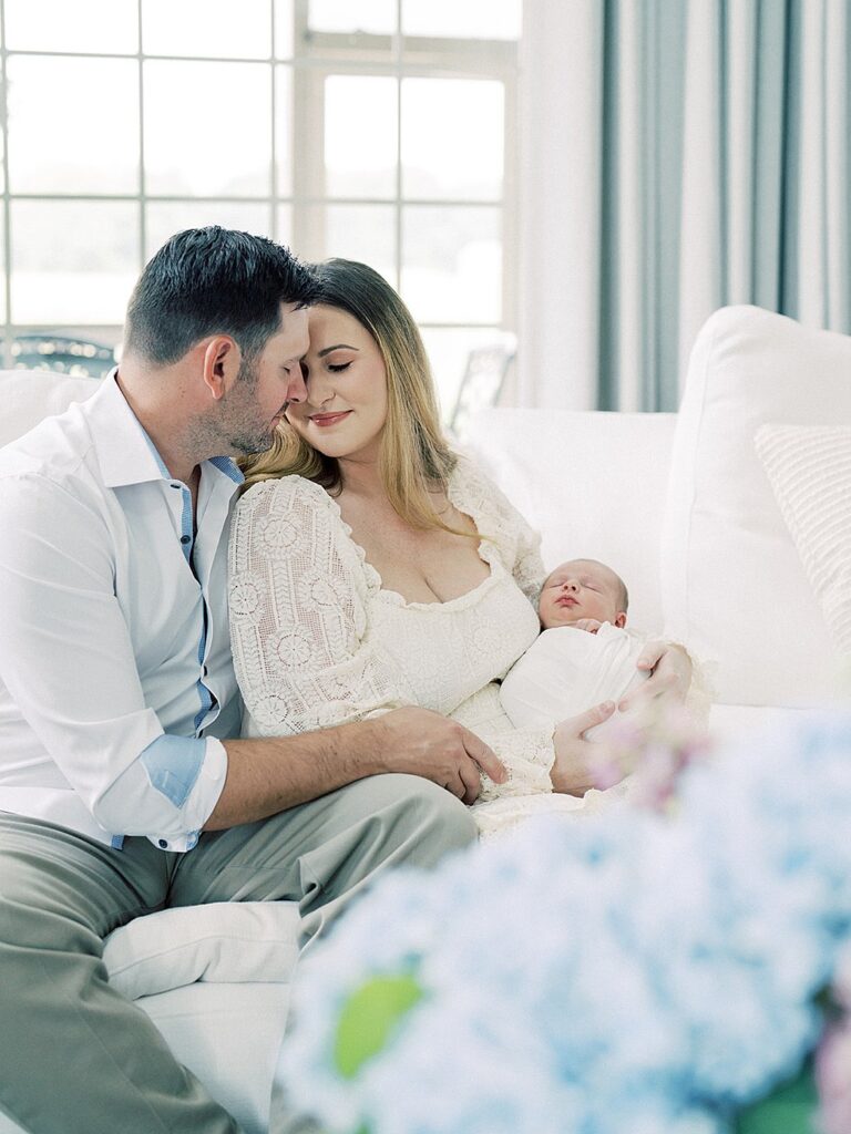 A Mother In White Lace Dress Leans Into Her Husband As They Sit On The Couch In Their Living Room Holding Their New Baby.