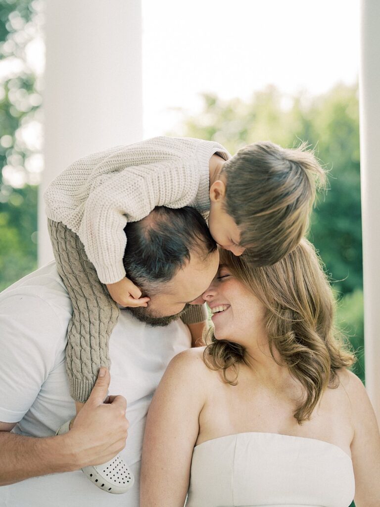 Little Boy On Dad's Shoulders Leans Down To Kiss His Mother On The Head.