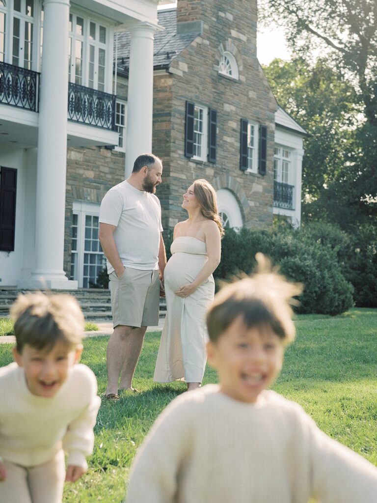 Two Boys Run Towards The Camera Out-Of-Focus As Their Parents Stand Behind Them Looking At One Another.