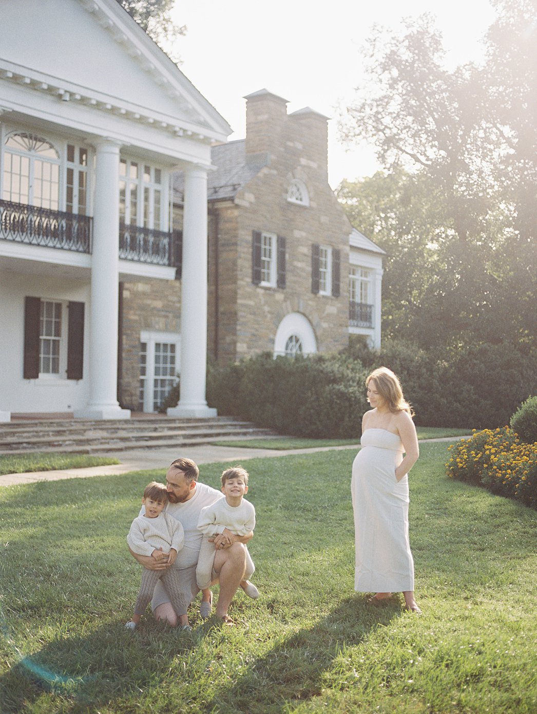 A Pregnant Mother Stands With Her Hands In Her Dress Pockets As She Watches Her Husband Crouch Down With Their Two Sons, Photographed By Rockville Md Family Photographer Marie Elizabeth Photography.