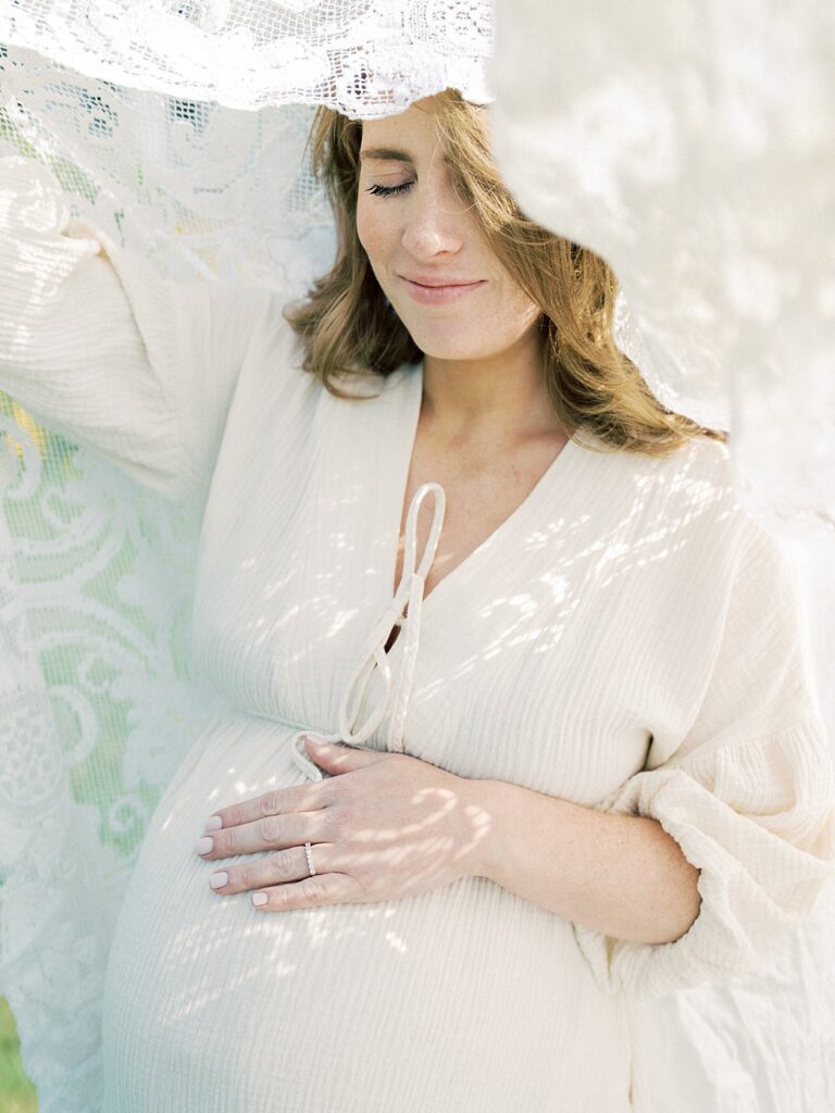 Red-Haired Mother Stands Under A Lace Blanket With Her Eyes Closed And One Hand On Her Belly, Photographed By One Of The Best Rockville Md Family Photographers, Marie Elizabeth Photography.