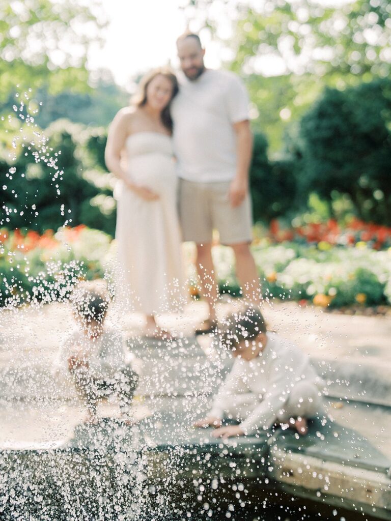 A Focused View Of The Fountain At Glenview Mansion While Two Little Boys And Their Parents Sit And Stand Out-Of-Focus Behind.