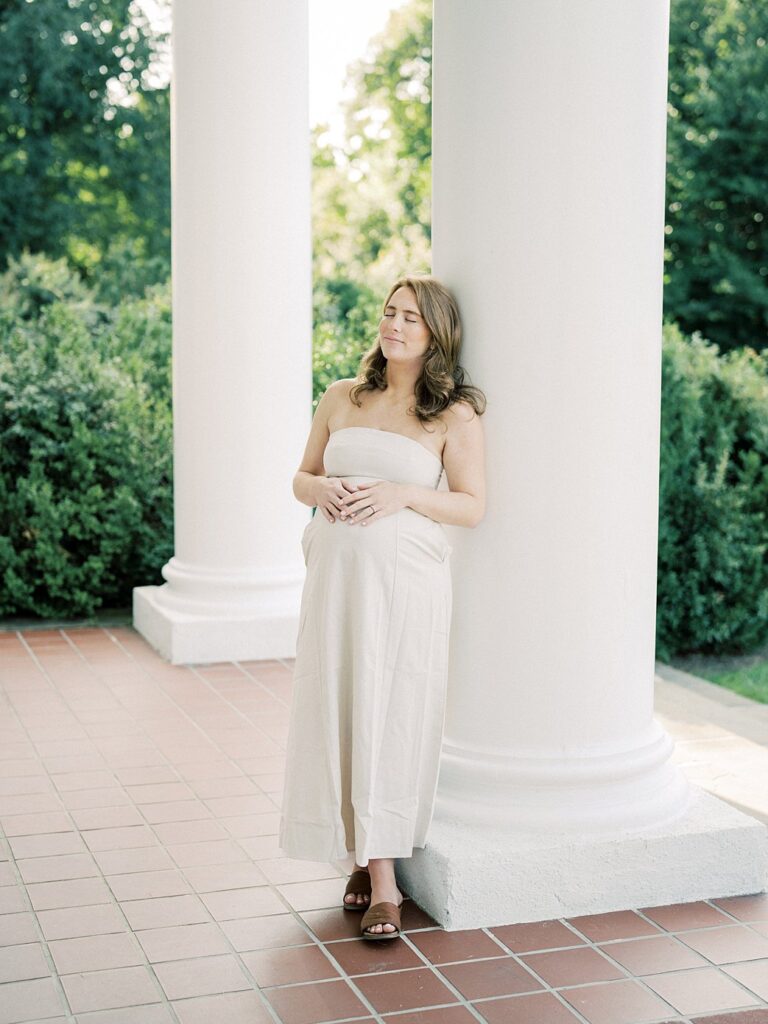 Pregnant Mom Leans Against A Pillar At Glenview Mansion In Rockville With Both Of Her Hands On Top Of Her Belly.