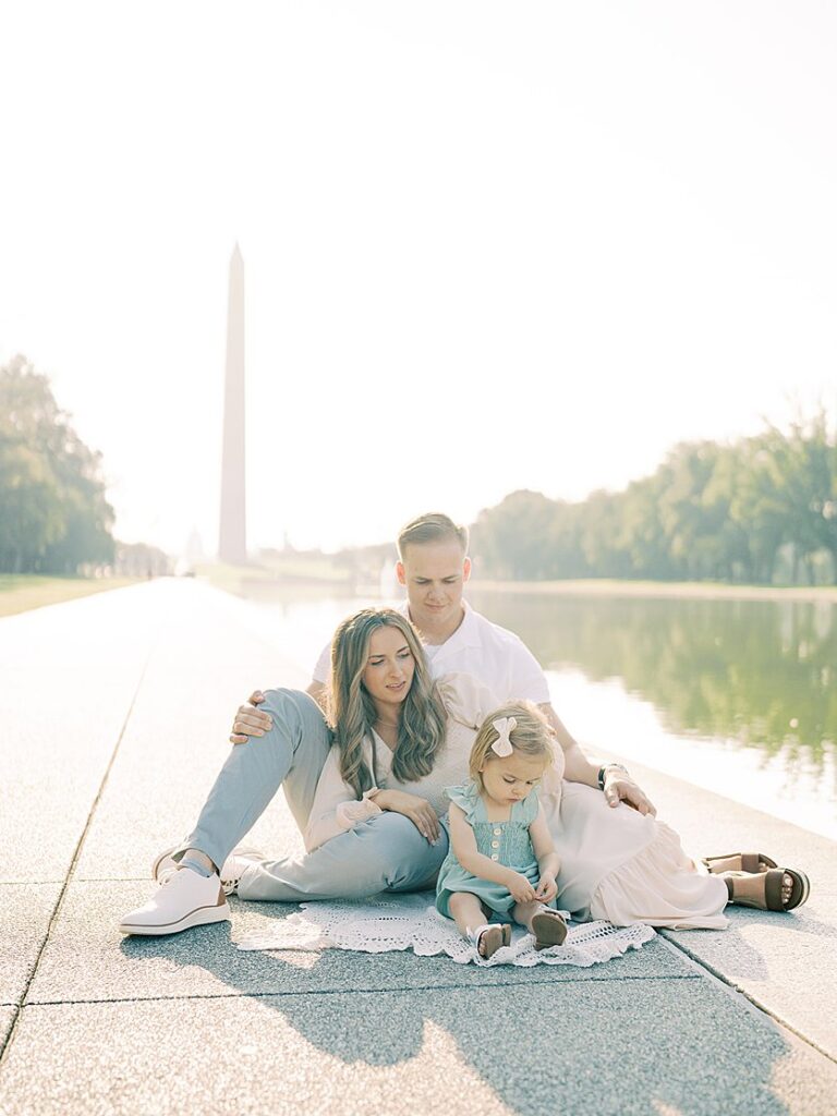 A Family Sits Along The Reflecting Pool In Dc With The Washington Monument In The Background.