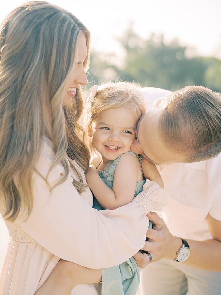 A Father Leans Down To Kiss His Little Girl On The Cheek As The Mother Holds Her During Their Constitution Gardens Family Photos.