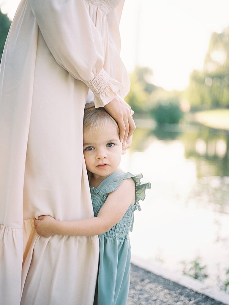 A Little Girl In A Sage Green Dress Hugs Her Mother And Looks At The Camera.