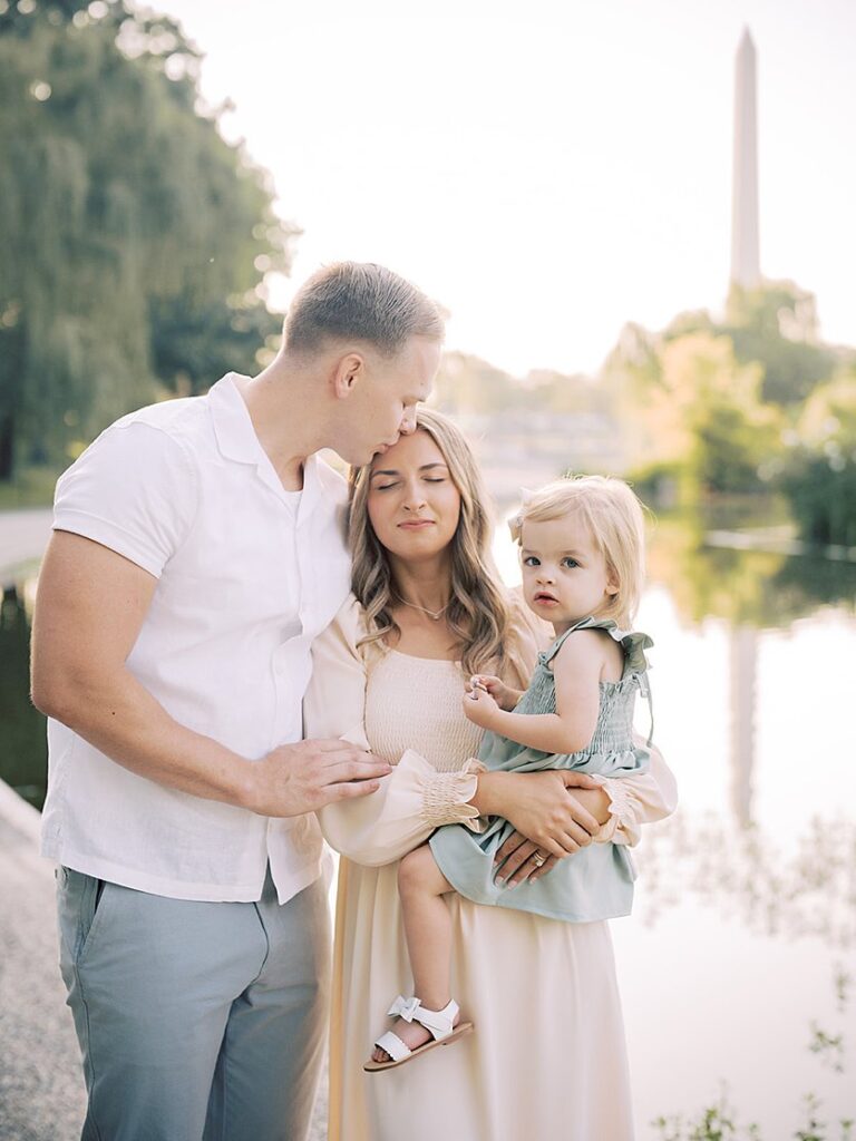 A Blonde Father Leans Over To Kiss His Wife On The Forehead As She Holds Their Toddler Daughter During Their Constitution Gardens Family Photos.