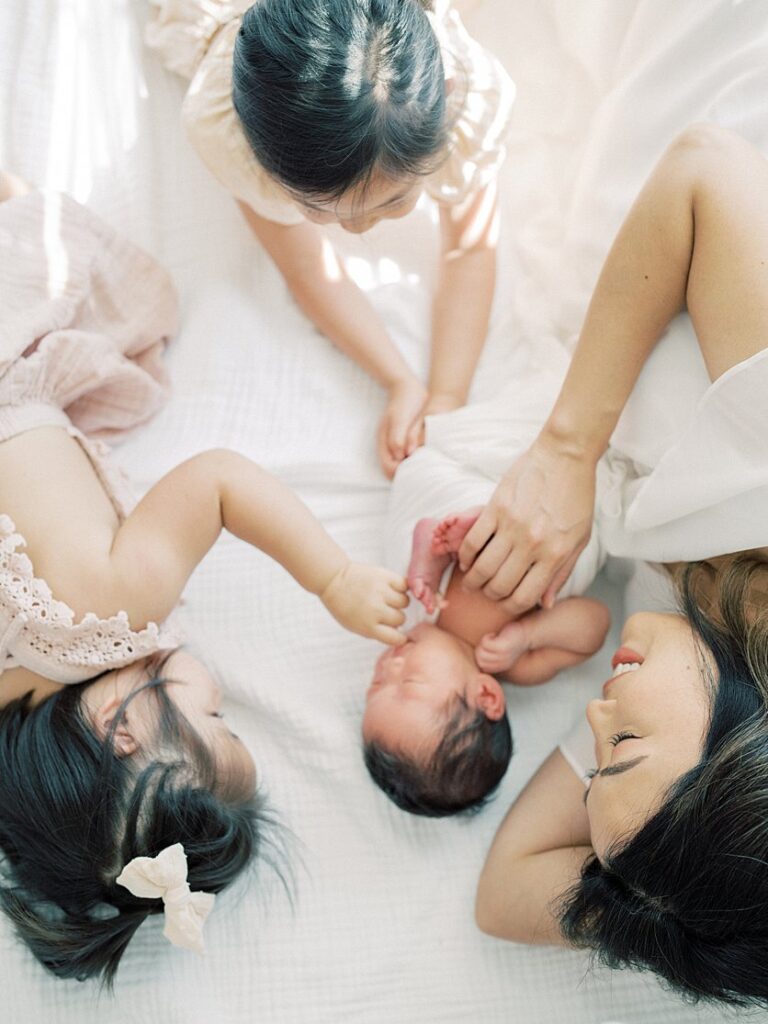 Mother Lays On Bed With Newborn Baby And Two Toddler Daughters During Their Falls Church Newborn Photography Session.