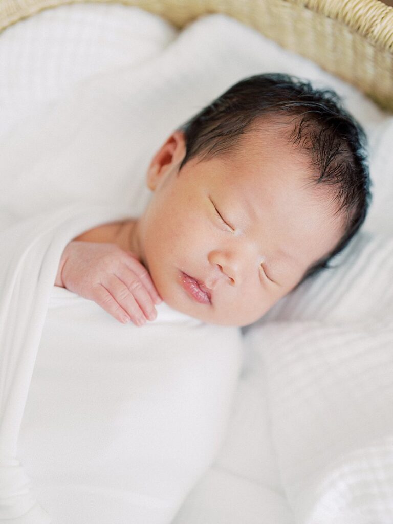 A Baby Swaddled In White Sleeps Peacefully In A Moses Basket With His Hand Resting On His Chest.