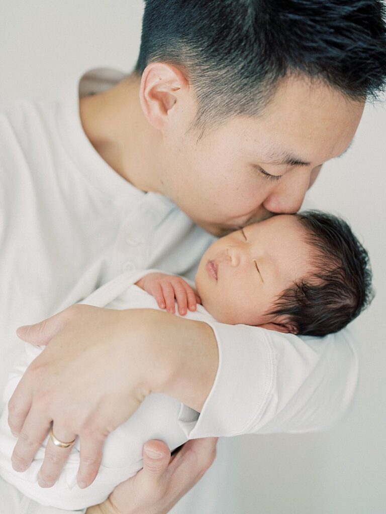 A Father Leans Over To Kiss His Baby In His Arms During His Falls Church Newborn Photography Session.