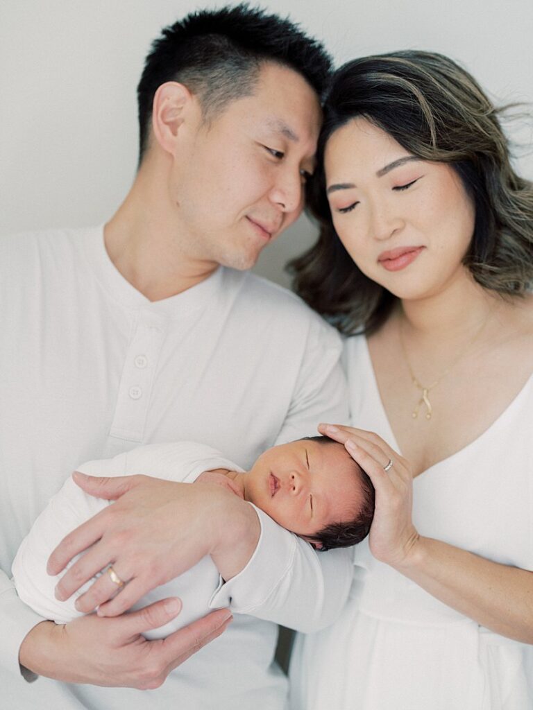 Mother And Father Lean Into One Another While Holding Their Swaddled Newborn Baby.