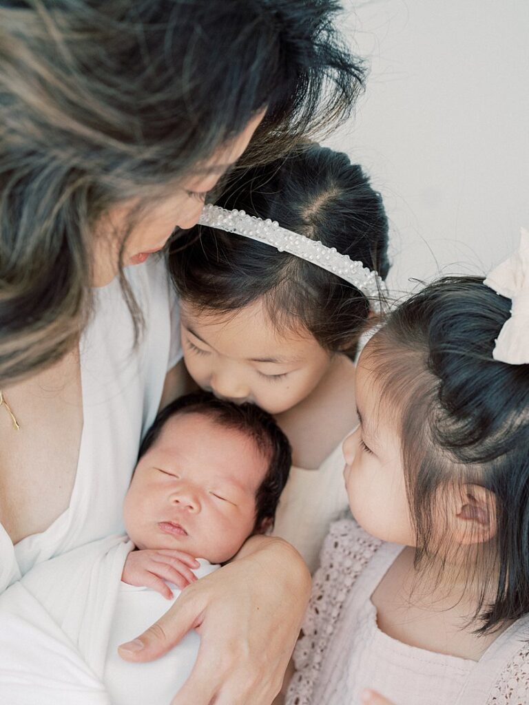 Two Toddler Girls Kiss And Admire Their Newborn Baby Brother As Mom Holds Baby.