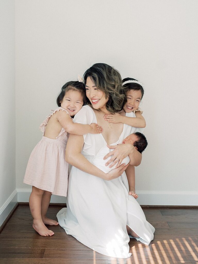 A Mother In White Dress Kneels As Her Two Toddler Daughters Hug Her As She Holds Her Baby.