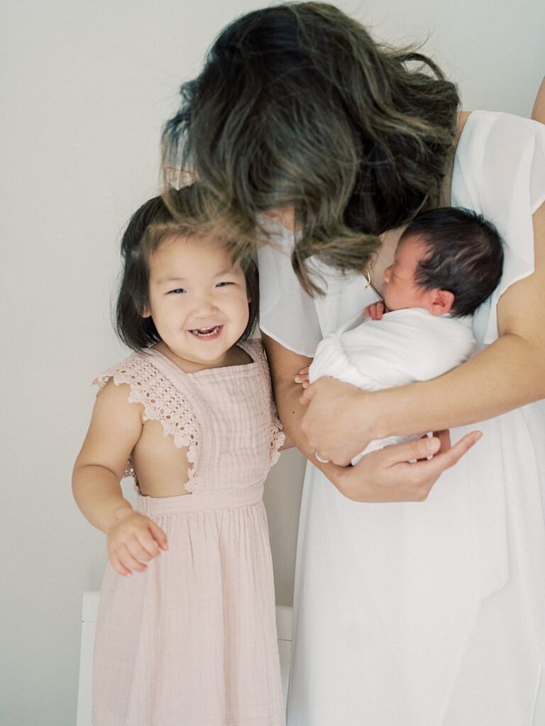 Toddler Girl In Pink Dress Smiles At The Camera As Her Mother Leans Down To Her While Holding Newborn.