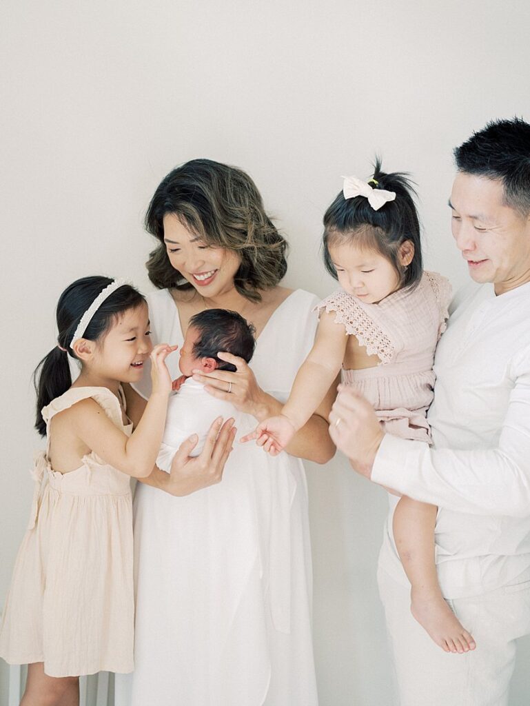 Mother Holds Newborn Baby Up To Her Toddler Daughter As Dad Holds Other Daughter Looking At Them.