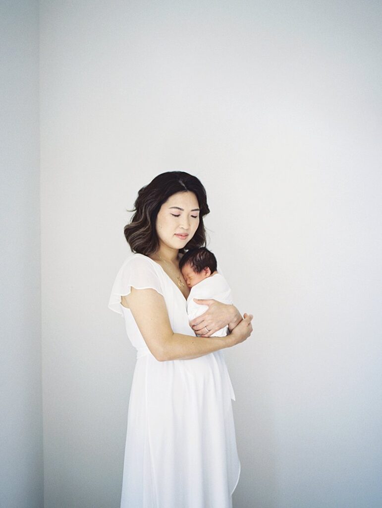 Asian Mother With Brown Hair In White Dress Stands Holding Her Newborn Baby Boy During Her Newborn Photography Session In Falls Church.