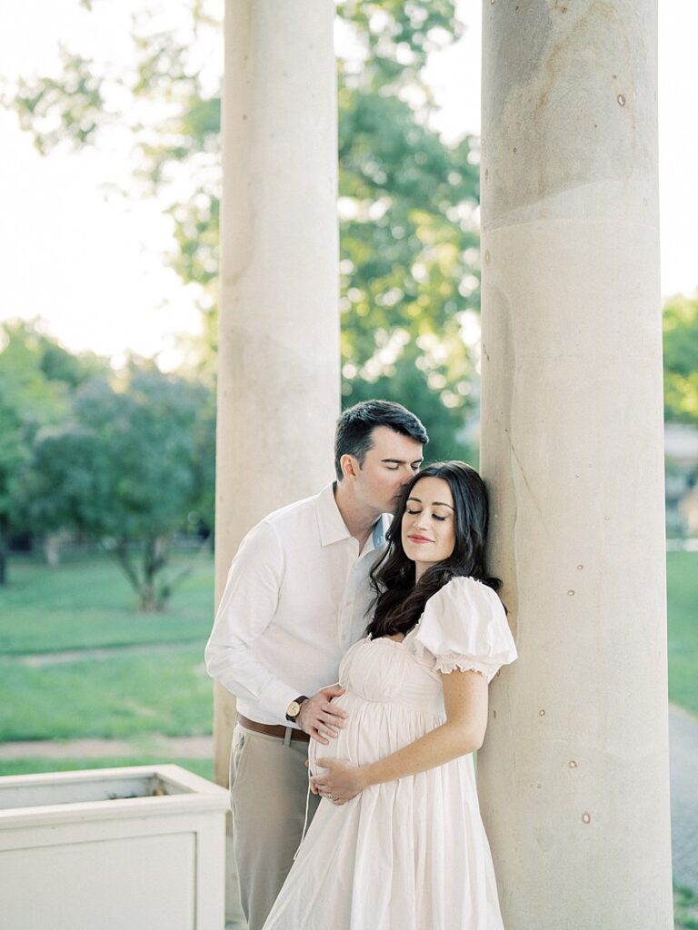 A Pregnant Mother Rests Against A Pillar While Her Husband Leans In To Kiss The Side Of Her Head.