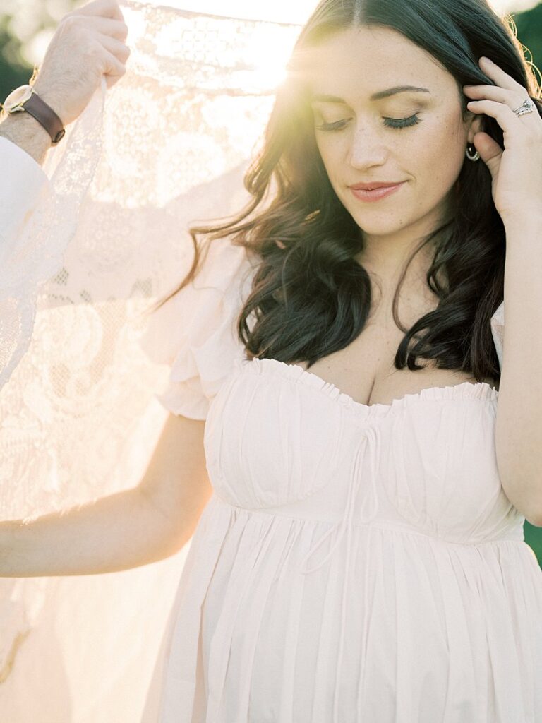 Pregnant Mom Stands With A Lace Blanket Held Up Behind Her During Their Maternity Session.