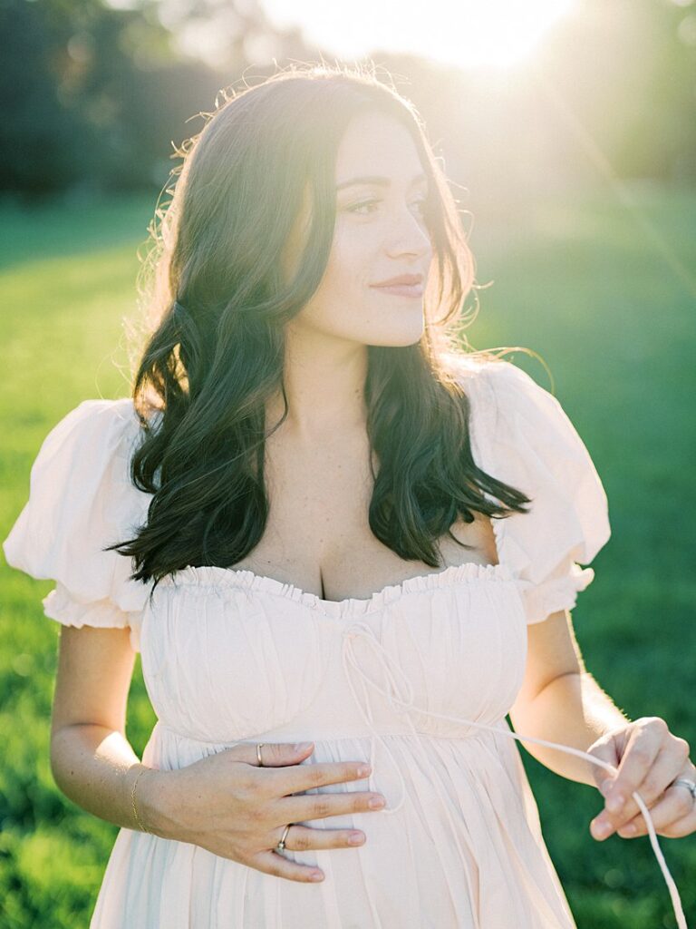 A Mother With Long Brown Hair Places One Hand On Her Pregnant Belly And One On Her Dress String And Looks To The Distance.