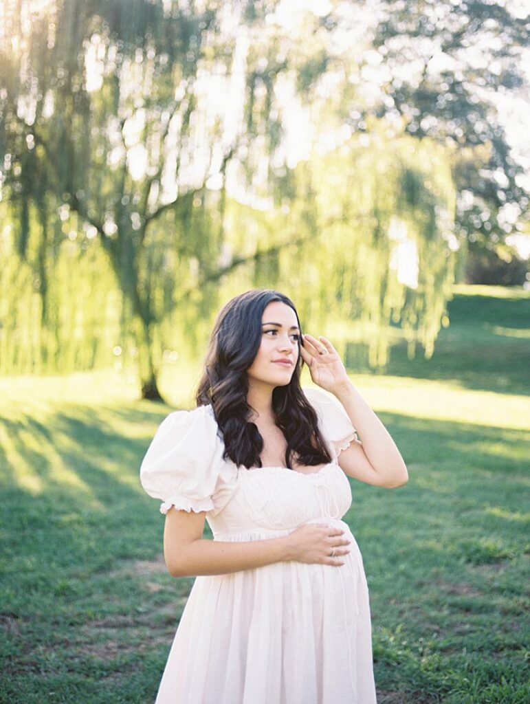 Pregnant Mother With Long Brown Hair Strokes Hair Aways From Her Face As She Gazes Off In The Distance While Standing In Front Of A Willow Tree.