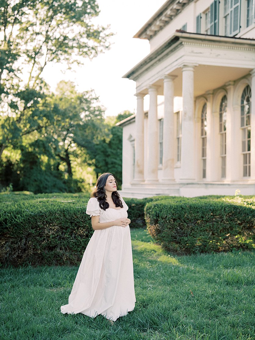 A Pregnant Mother In A Long Pink Gown Stands In Front Of Riversdale Manor In Maryland During Her Maternity Session.