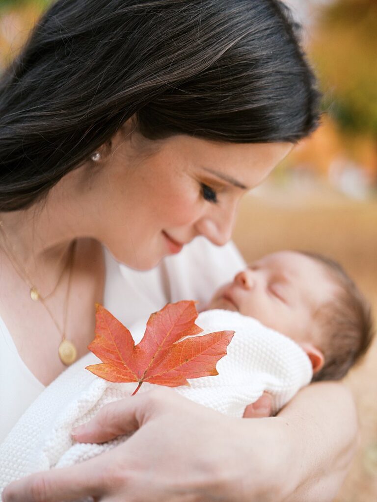 A Mother Holds Her Newborn Baby Up To Her Nose While Grasping An Orange Maple Leaf During Their Rockville Newborn Photography Session.