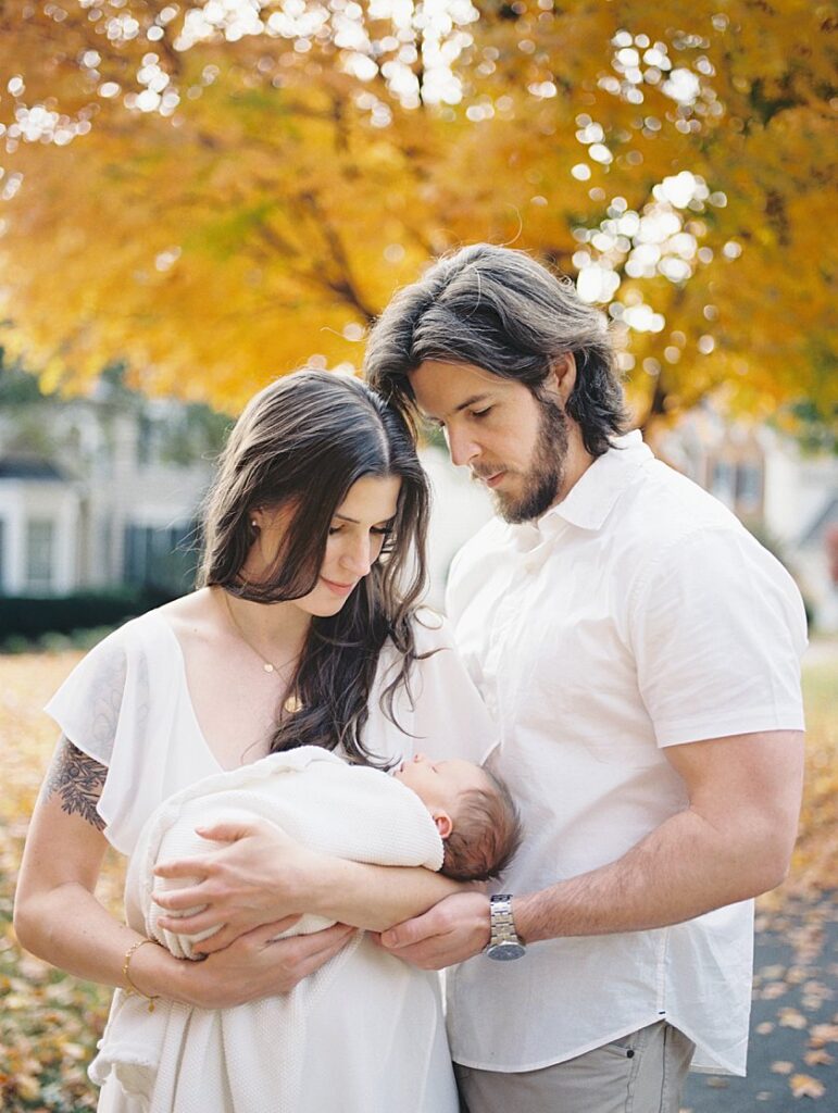 A New Father And Mother Stand Outside In Front Of An Orange Maple Tree During Their Rockville Newborn Photography Session.